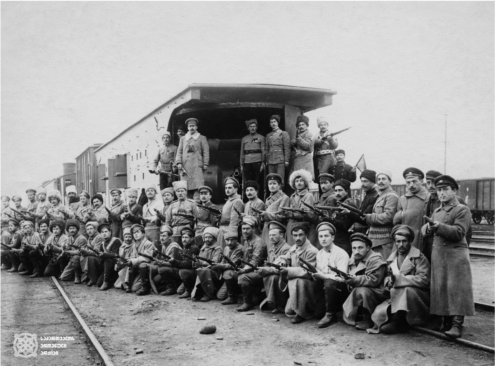 სახალხო გვარდიის ჯავშანმატარებელთა რაზმის წევრები, ჯავშანმატარებელი N1 „რესპუბლიკელის“ ფონზე. ზედა რიგში მაუზერით ხელში და ბინოკლით რაზმის მეთაური, ვლადიმერ გოგუაძე. 1918-1921 წლები
<br>
Members of the division of armored train, on the background of armored train N1 “Respublikeli”. In the upper row is commander of the division Valodia Goguadze. 1918-1921