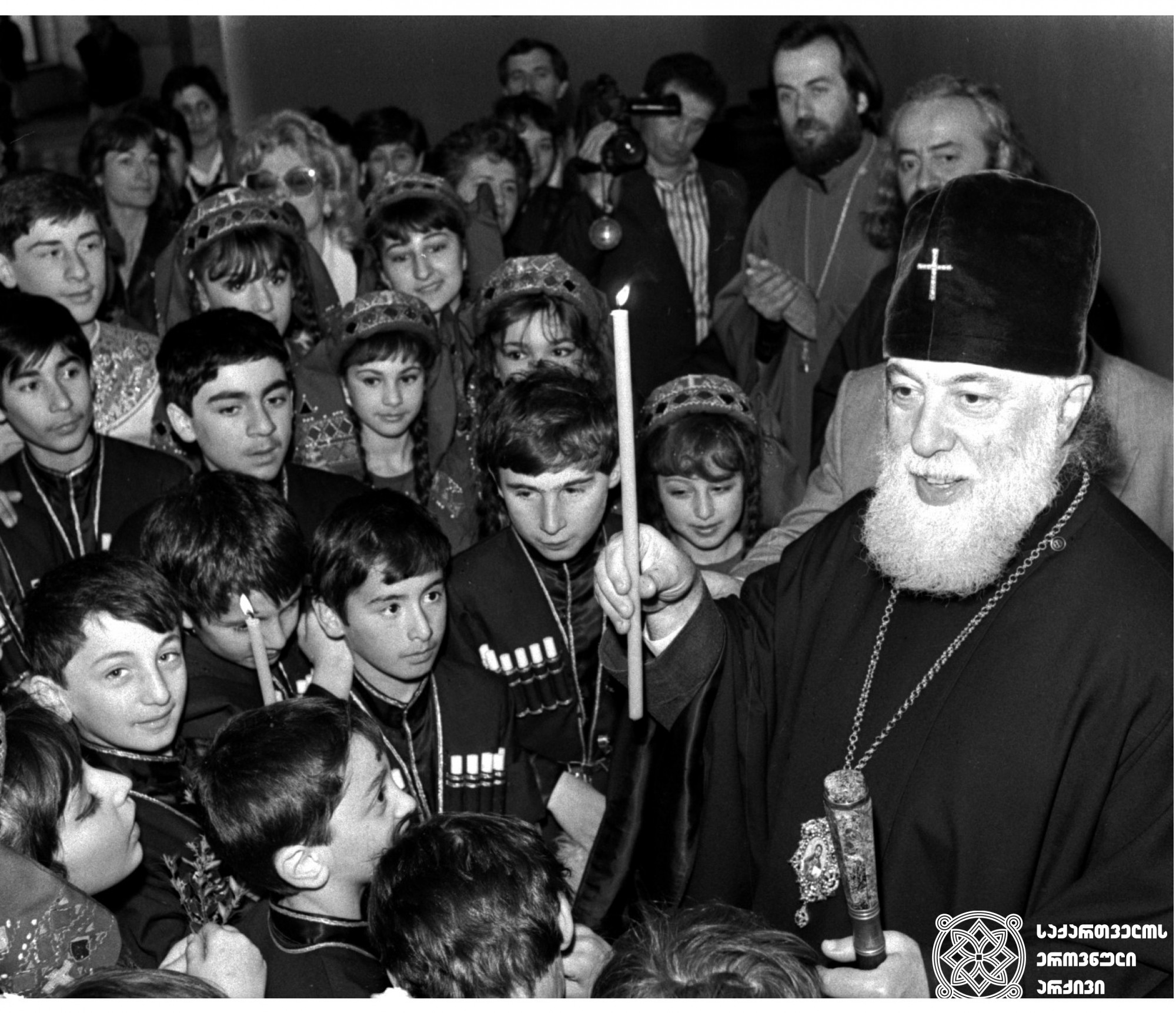 ილია II სრულიად საქართველოს კათოლიკოს-პატრიარქი ბავშვებთან ერთად ბანკის კურთხევისას <br>
თბილისი, 1992 წელი <br>
Ilia II, Catholicos-Patriarch of All Georgia, with children during the blessing of a bank  <br>
Tbilisi, 1992