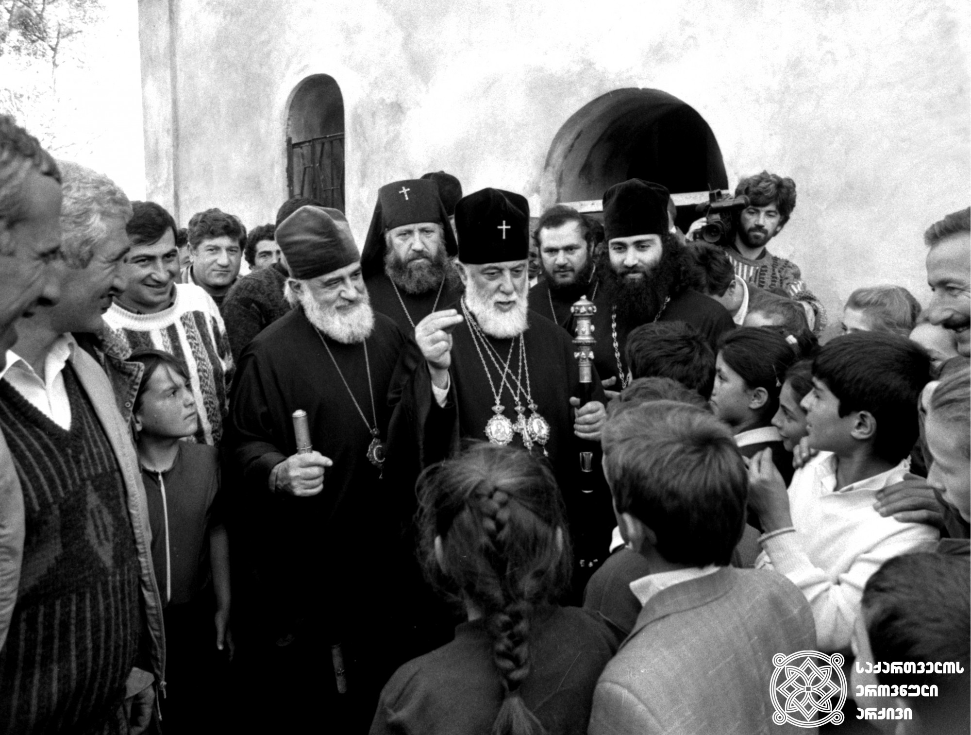 ილია II სრულიად საქართველოს კათოლიკოს-პატრიარქი პირჯვრის წერას ასწავლის სოხუმელ ბავშვებს <br>
პატრიარქისგან მარჯვნივ: მიტროპოლიტი დავითი (ჭკადუა), უკან: მიტროპოლიტი ამბროსი (ქათამაძე), მარცხნივ: მამა ელიზბარი (ელიზბარაშვილი) <br>
სოხუმის საკათედრო ტაძრის წინ, 1991 წელი <br>
Ilia II, Catholicos-Patriarch of All Georgia teaching children from Sokhumi how to make the Sign of the Cross <br>
To the right of the Patriarch: Metropolitan Davit (Chkadua); Backwards: Metropolitan Ambrosi (Katamadze); To the left: Father Elizbar (Elizbarashvili) <br>
In front of the Sukhumi Cathedral, 1991
